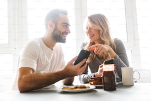 Image d’un jeune couple amoureux incroyable assis à la cuisine prendre un petit déjeuner en utilisant des téléphones portables.