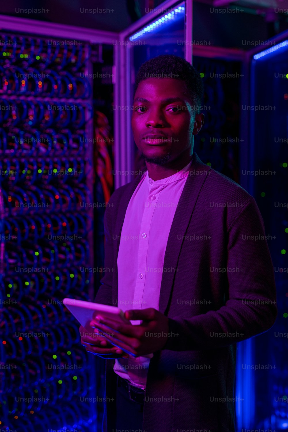 Portrait of serious young Black network security engineer in cardigan standing with tablet against server rack cabinet