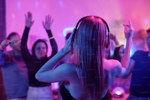 Back view of young woman with long blond hair touching headphones while standing in front of crowd of excited friends dancing at party