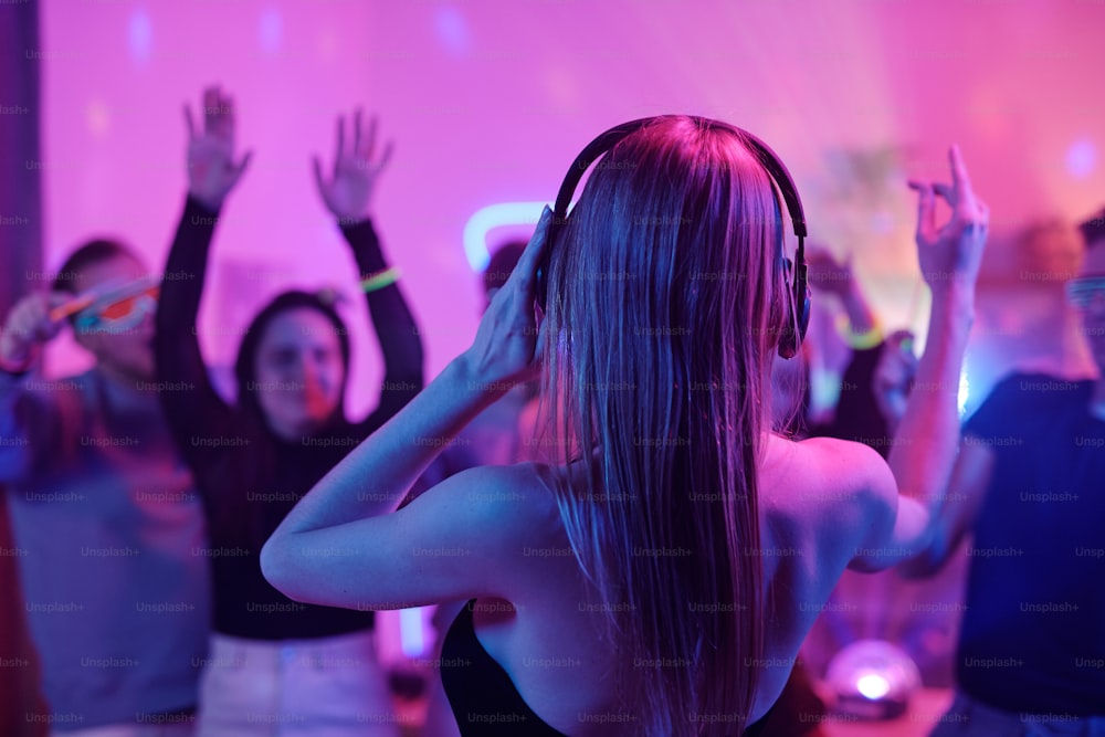 Back view of young woman with long blond hair touching headphones while standing in front of crowd of excited friends dancing at party