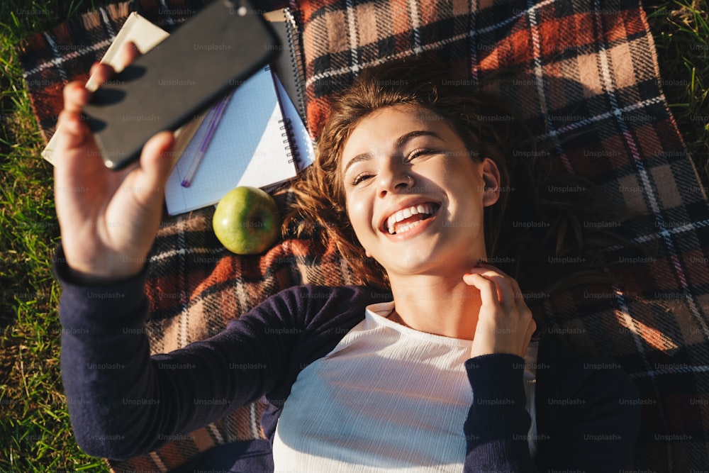 Vista superior de uma jovem alegre deitada em um cobertor no parque, relaxando, tirando uma selfie