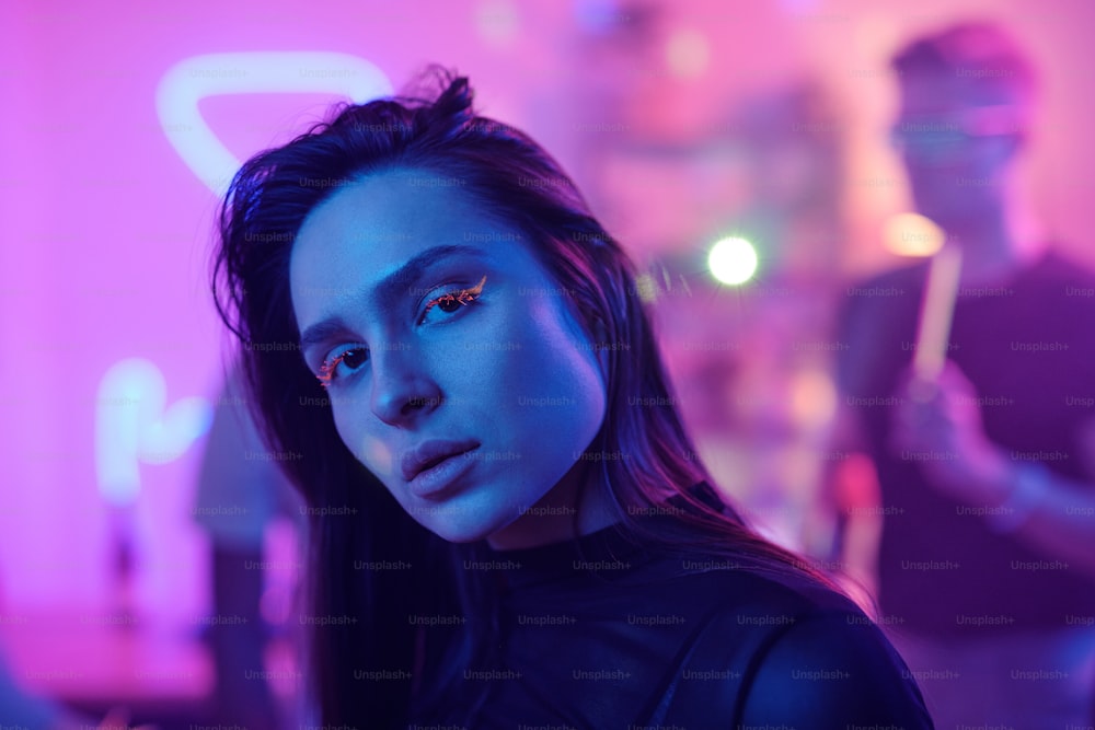 Young woman with glamorous neon makeup looking at you while standing in front of camera against her friends dancing and enjoying party