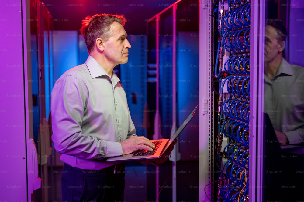 Ingegnere di rete caucasico maturo concentrato in camicia in piedi all'armadio del server aperto ed esegue il controllo di sicurezza utilizzando il laptop