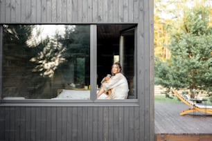 Mujer joven descansando en una hermosa casa de campo u hotel, sentada en el alféizar de la ventana disfrutando de una hermosa vista sobre el bosque de pinos. Vista desde el exterior. Concepto de soledad y recreación en la naturaleza