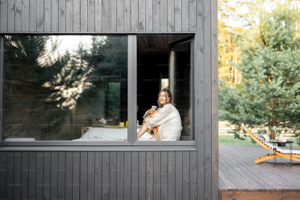 Jeune femme se reposant dans une belle maison de campagne ou un hôtel, assise sur le rebord de la fenêtre bénéficiant d’une vue magnifique sur la forêt de pins. Vue de l’extérieur. Concept de solitude et de loisirs sur la nature