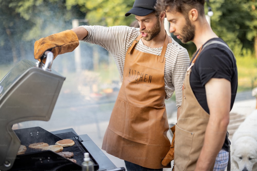 두 명의 잘생긴 남자 친구가 자연 뒤뜰의 가스 그릴에서 햄버거를 위해 고기와 빵을 굽는 동안 즐거운 시간을 보내고 있습니다