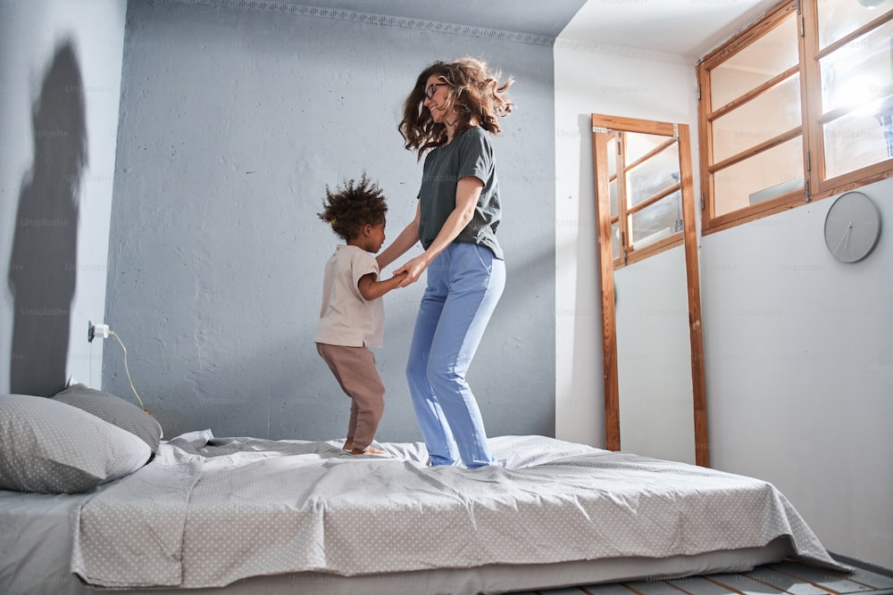 Full length view of the happy mother jumping at the bed with her multiracial daughter while feeling great at the morning at their apartments. Stock photo