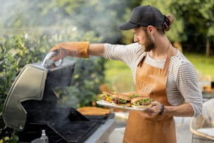 Handsome man in cap and apron making burgers on a grill at backyard. Cooking outdoors and american lifestyle concept