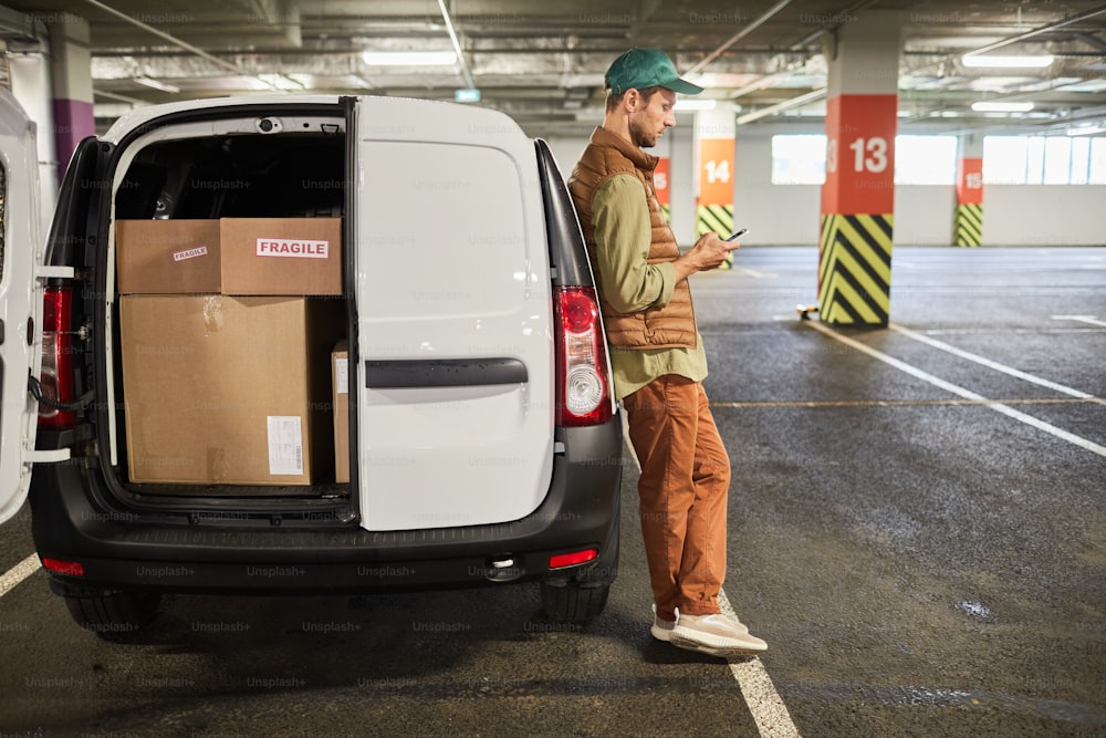 Retrato de corpo inteiro do entregador do sexo masculino em pé pela van cheia de caixas no estacionamento, espaço de cópia