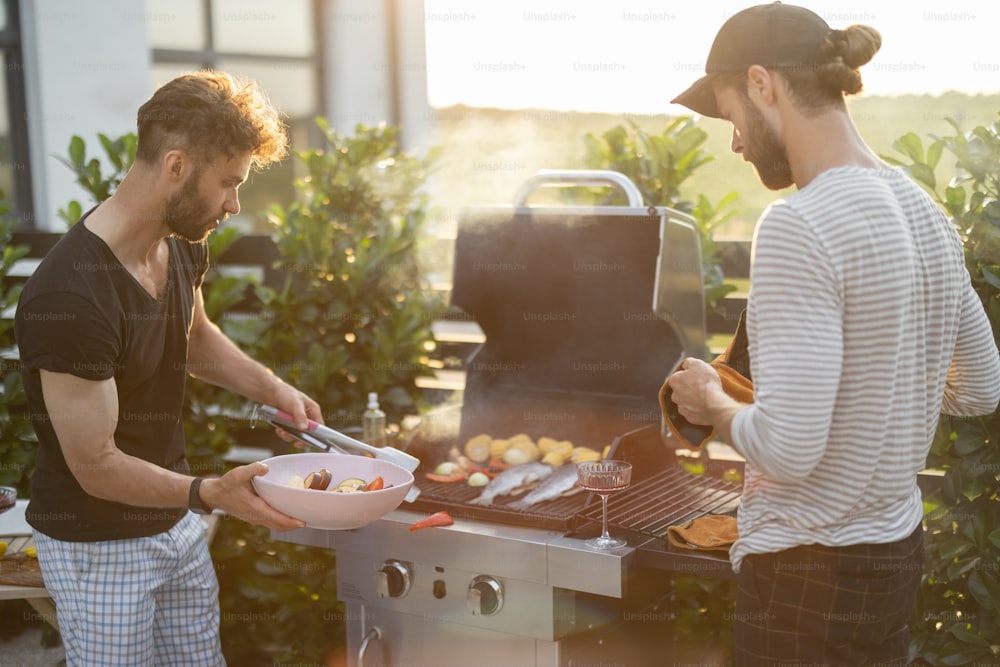 Two caucasian guys cooking vegetables and fish on a modern gas grill at backyard on a sunset. Healthy eating on the open air, male friendship