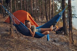 Female lying in hammock with her legs and looking at camera with orange tent and campfire in background