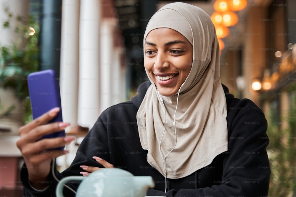 Vista de retrato de cintura hacia arriba de la mujer islámica relajada está sentada a la mesa con un teléfono inteligente al aire libre y charlando con alguien a través de una videollamada mientras toma café o té. Foto de archivo