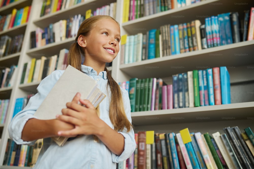 Angle bas d’une belle écolière souriante avec une pile de livres regardant au loin