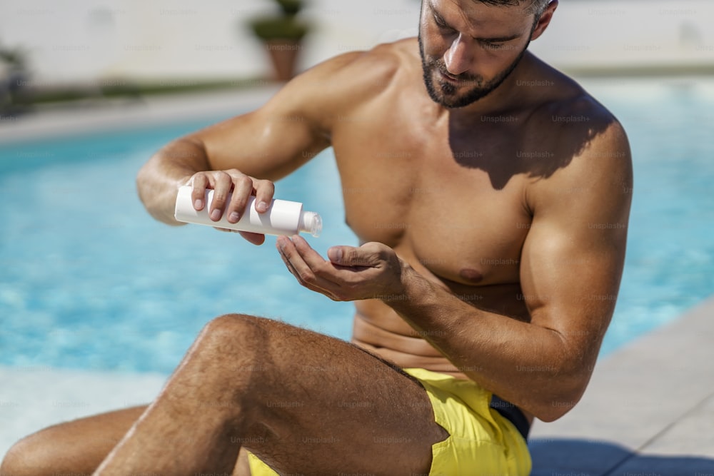 Sonnenschutz, Kosmetik und Cremes. Ein sexy Mann trägt Sonnencreme und Sonnencreme auf, während er in einem Badeanzug am Pool sitzt. Hautschutz und Hautpflege vor UV-Strahlung, lieben Sie Ihre Haut und Ihren Körper