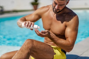 Sonnenschutz, Kosmetik und Cremes. Ein sexy Mann trägt Sonnencreme und Sonnencreme auf, während er in einem Badeanzug am Pool sitzt. Hautschutz und Hautpflege vor UV-Strahlung, lieben Sie Ihre Haut und Ihren Körper