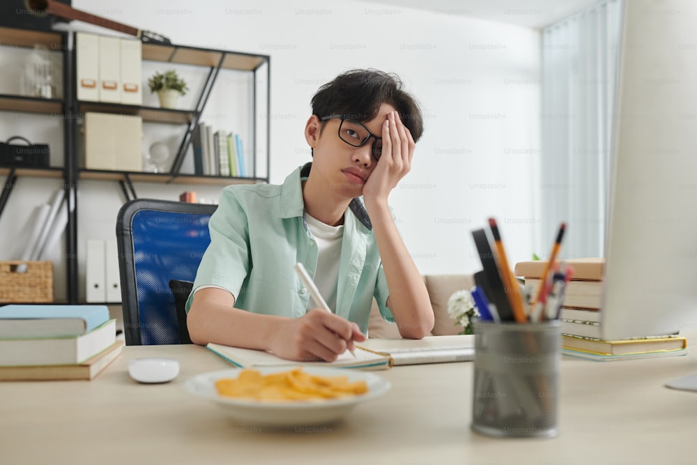 Teenage boy tired of doing homework at his desk at home, he is looking at computer screen and coverig face with hand