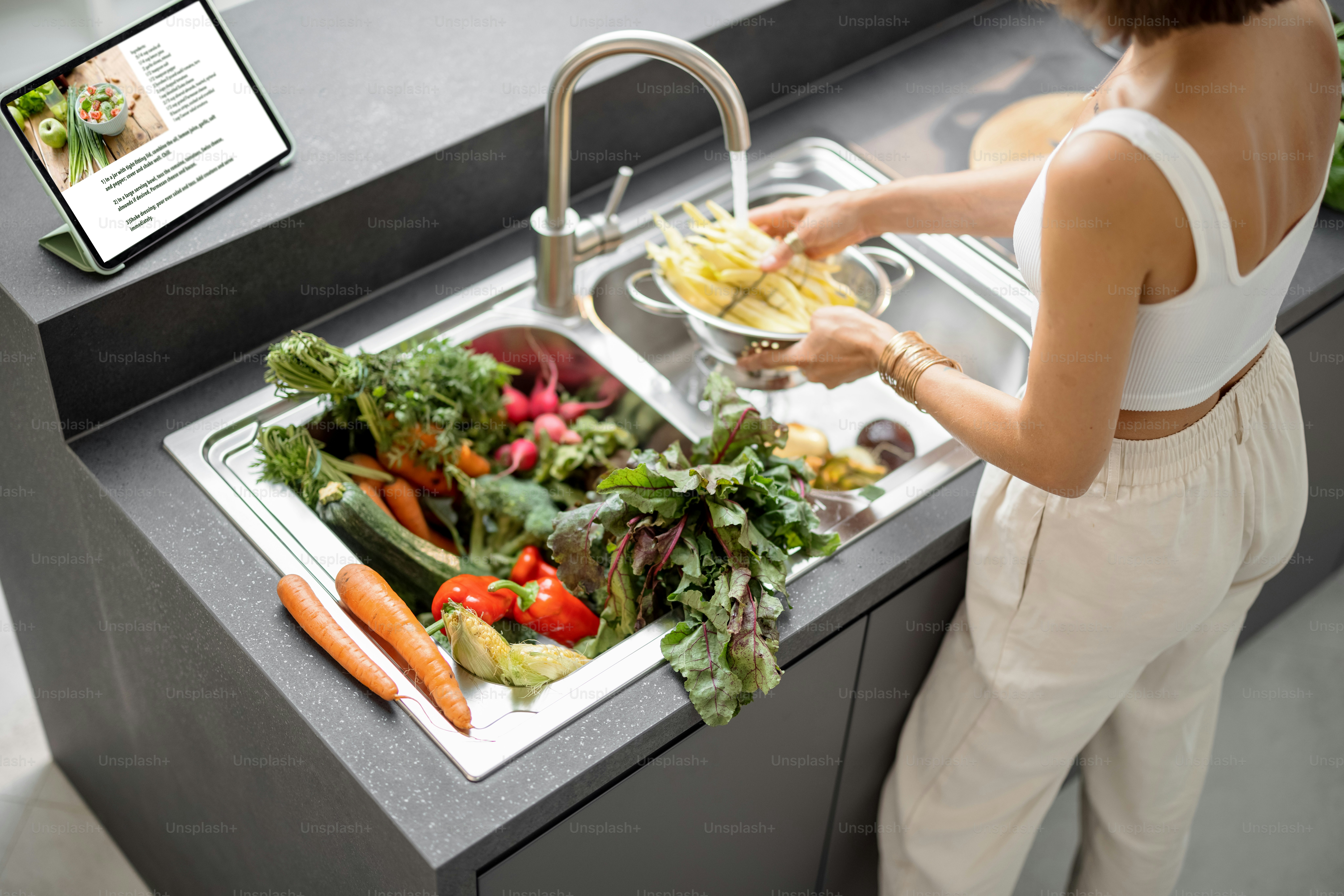 Giovane donna che cucina con verdure fresche e verdure che guardano la ricetta sul tablet digitale nella cucina moderna. Ingredienti biologici freschi, igiene e concetto di alimentazione sana. Vista dall'alto