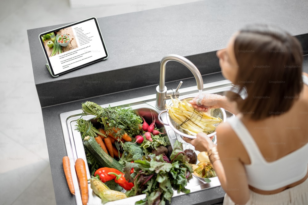 Mujer joven cocinando con verduras frescas y verduras mirando la receta en la tableta digital en la cocina moderna. Ingredientes orgánicos frescos, higiene y concepto de alimentación saludable. Vista desde arriba