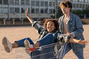 Happy African girl outstretching arms while sitting in shopping cart pushed by her boyfriend