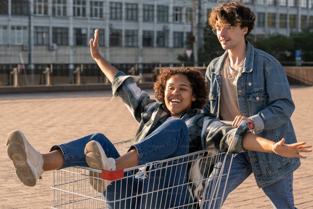 Happy African girl outstretching arms while sitting in shopping cart pushed by her boyfriend