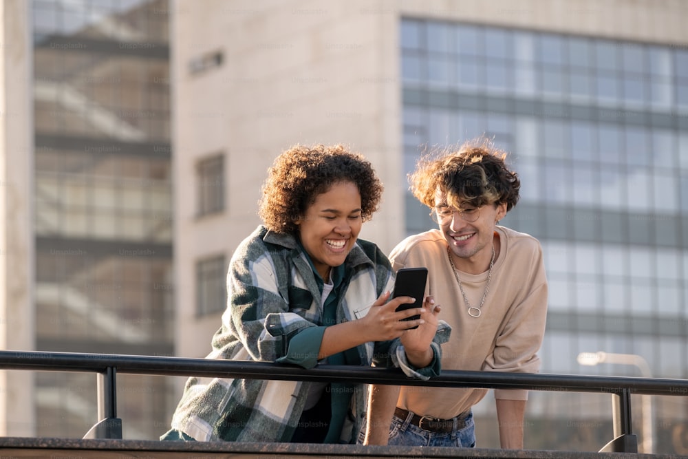 Lachendes gemischtrassiges Teenager-Mädchen mit Smartphone, das ihrem Freund etwas Lustiges zeigt