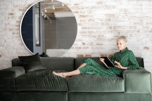 Stilvolle Frau im grünen Kleid mit digitalem Tablet, während sie entspannt auf dem bequemen Sofa zu Hause sitzt. Freizeit mit digitalem Endgerät oder Working-Home-Konzept