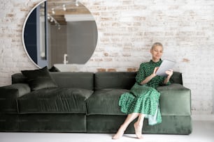 Stylish woman in green dress using digital tablet while sitting relaxed on the comfortable sofa at home. Leisure time with a digital device or working home concept