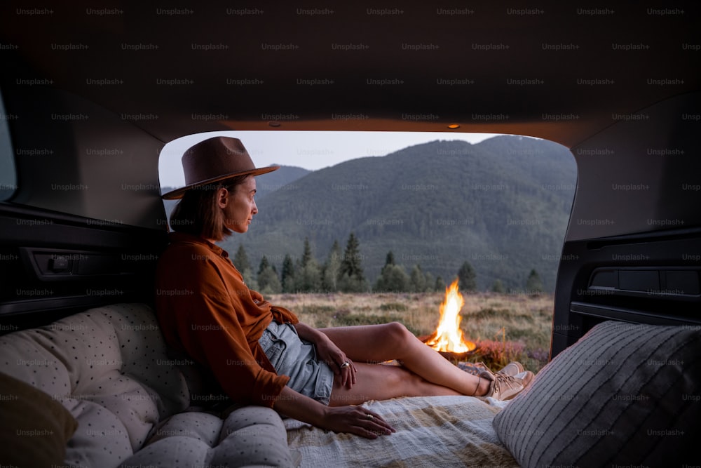 La donna ama il falò, sedersi e rilassarsi nel bagagliaio del veicolo con cuscini. Viaggiare in auto, romanticismo al picnic in montagna