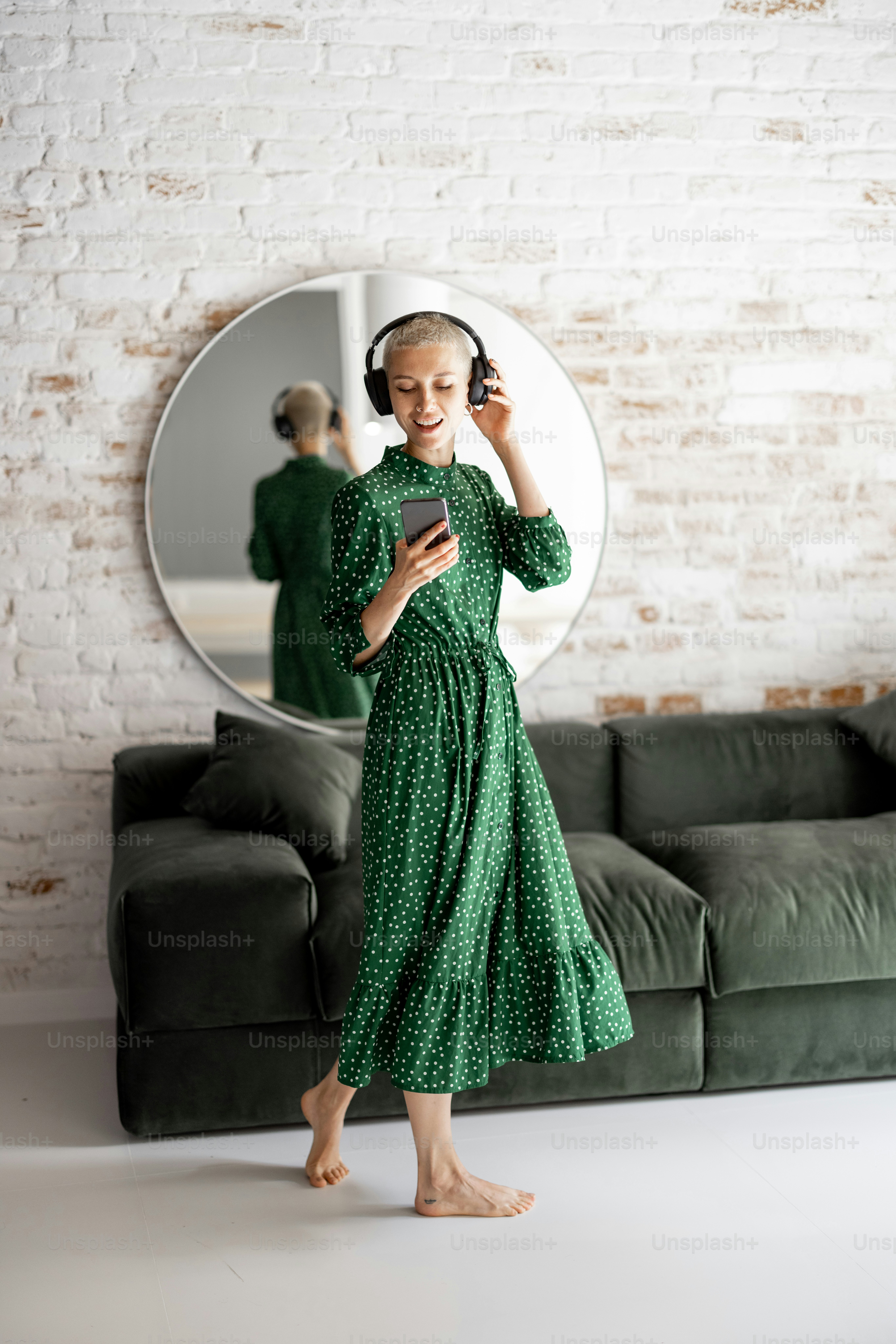 Stylish woman in green dress enjoys the music with headphones and cell phone dancing in the living room at home
