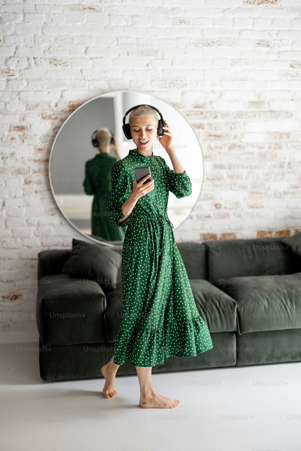 초록색 드레스를 입은 세련된 여성은 집 거실에서 헤드폰과 휴대폰 춤을 추며 음악을 즐긴다