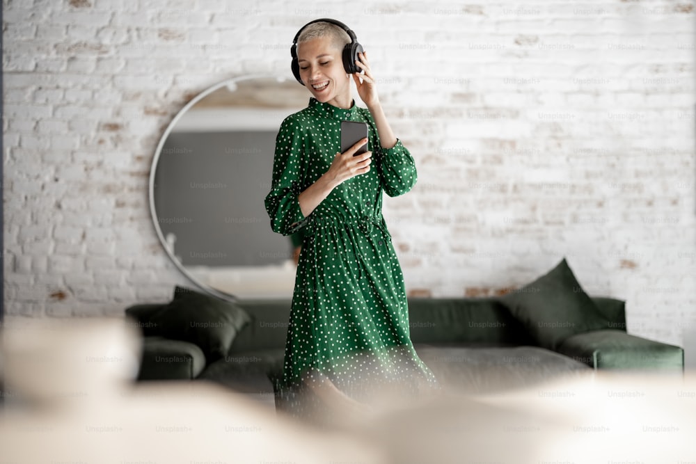 Mujer elegante con vestido verde disfruta de la música con auriculares y teléfono celular bailando en la sala de estar de su casa. Concepto de confort de vida y tiempo libre