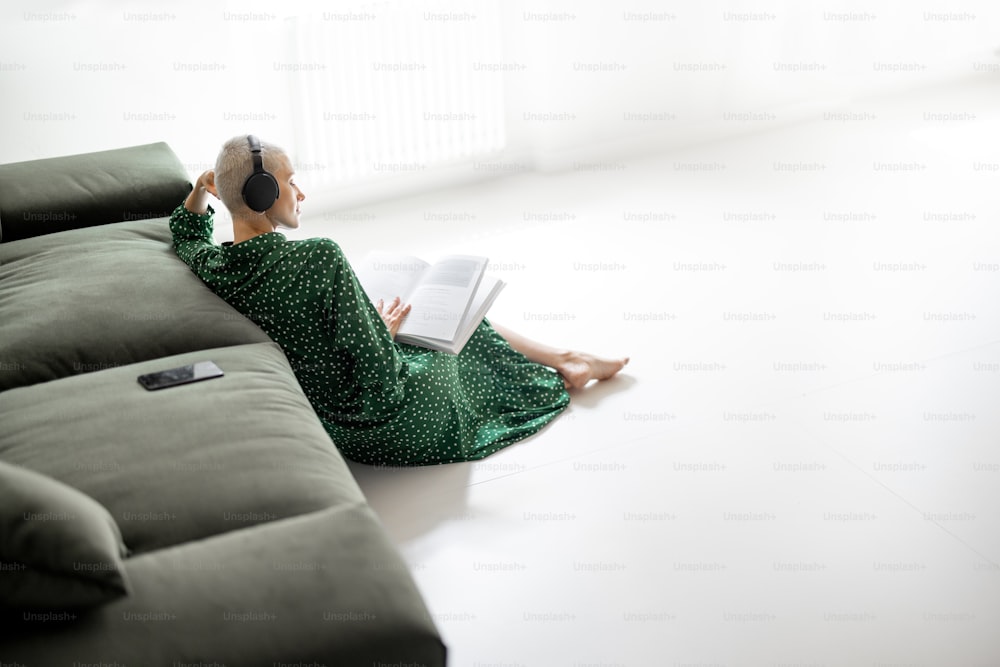 Mujer elegante con vestido verde leyendo el libro y escuchando la música, mientras está sentada en el suelo cerca del sofá de su casa. Tiempo libre y comodidad viviendo en casa