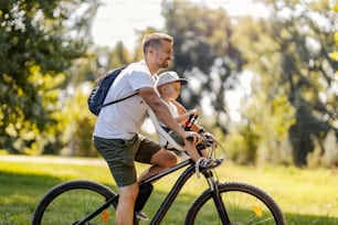 가족 사이클링. 아버지와 아들은 화창한 여름날 푸른 공원에서 자전거를 타다가 휴식을 취합니다. 모자를 쓴 유아가 자전거 바구니에 앉아 있고 아빠는 그 옆에 서 있습니다. 측면도