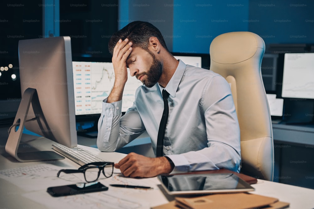 Frustrated young man in shirt and tie having headache while staying late in the office