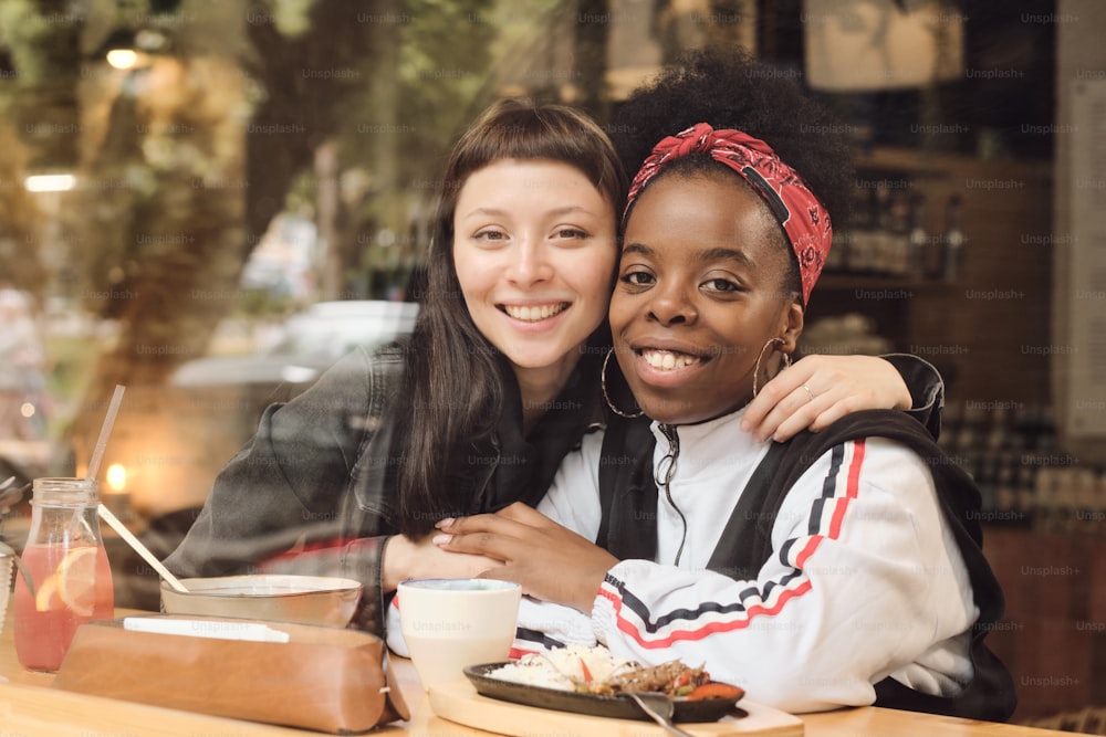 Two young gorgeous girlfriends sitting by served table in embrace during lunch