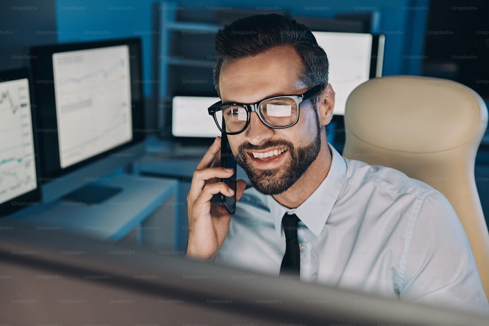 Jeune homme heureux parlant au téléphone portable et regardant l’ordinateur tout en restant tard au bureau