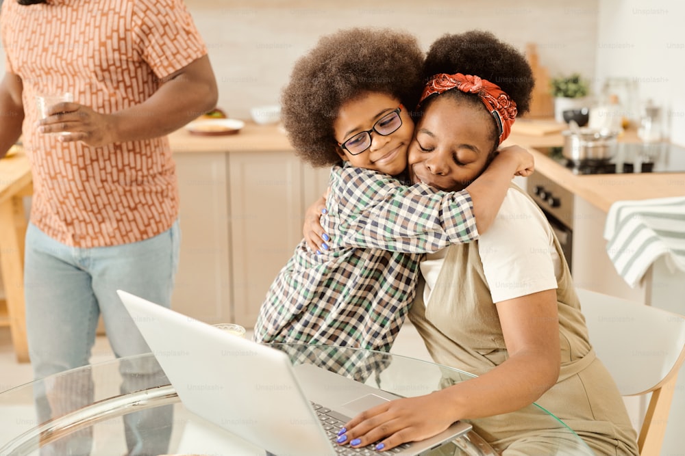 Cute little boy embracing his mom networking in front of laptop by kitchen table
