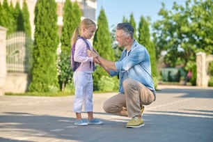 Entendimiento mutuo. Atento y atento hombre de mediana edad agazapado cerca de su pequeña hija con mochila caminando por la calle verde en una tarde