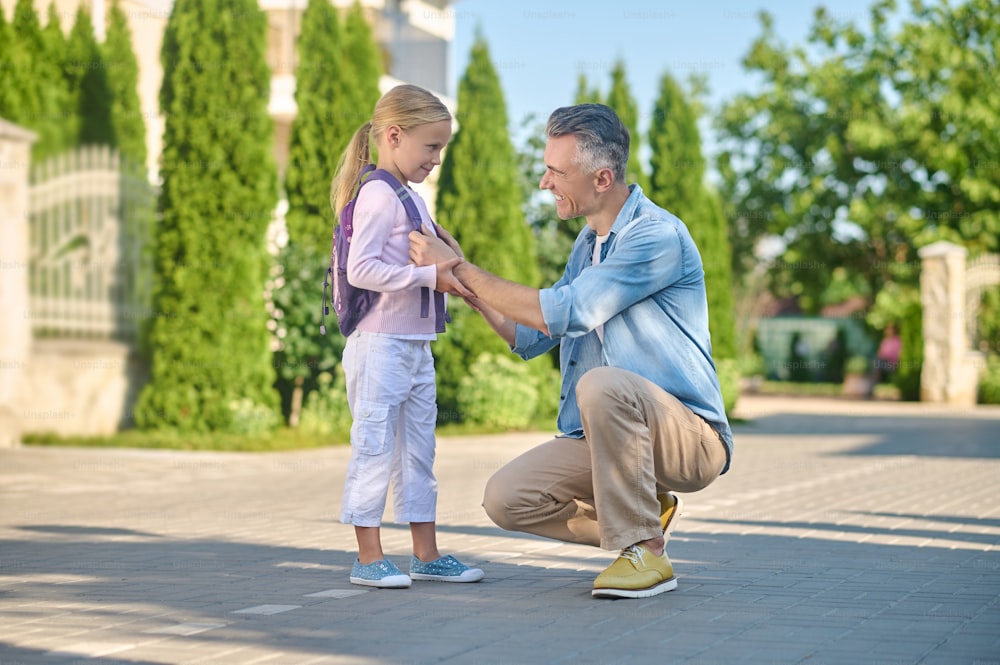 Compréhension. Homme d’âge moyen attentionné et attentionné accroupi près de sa petite fille avec sac à dos marchant dans la rue verte l’après-midi
