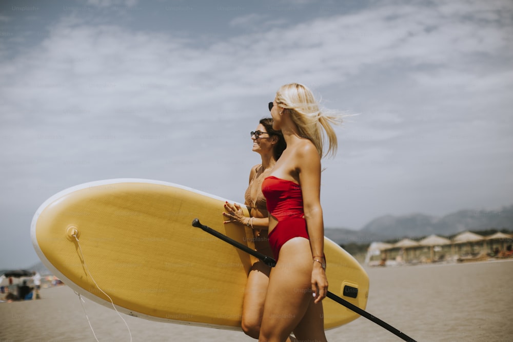Zwei hübsche junge Frauen mit Paddelbrett am Strand an einem Sommertag