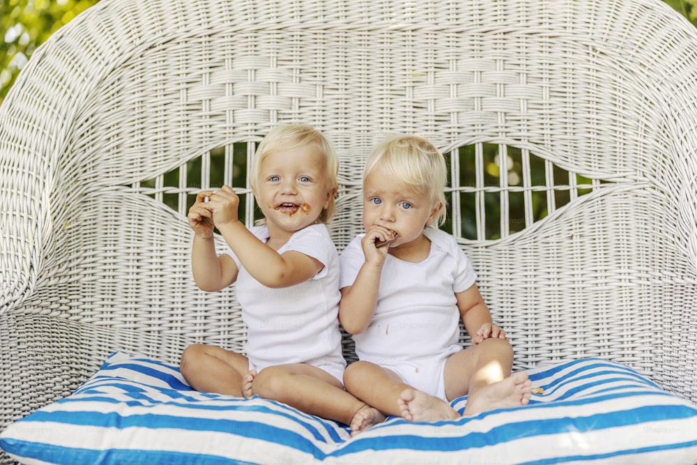 幼児の双子は庭の籐の椅子でクッキーを食べています。白い赤ちゃんの体を着たかわいい赤ちゃんがケーキを手に持っています。明るい髪、青い目、幼児の赤ちゃん、平和な子供時代