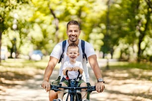 Father and son bonding time in nature. A little boy is sitting in a bicycle basket while his father rides a bicycle through the woods. Beautiful sunny day in nature to hanging out with family. Smiling