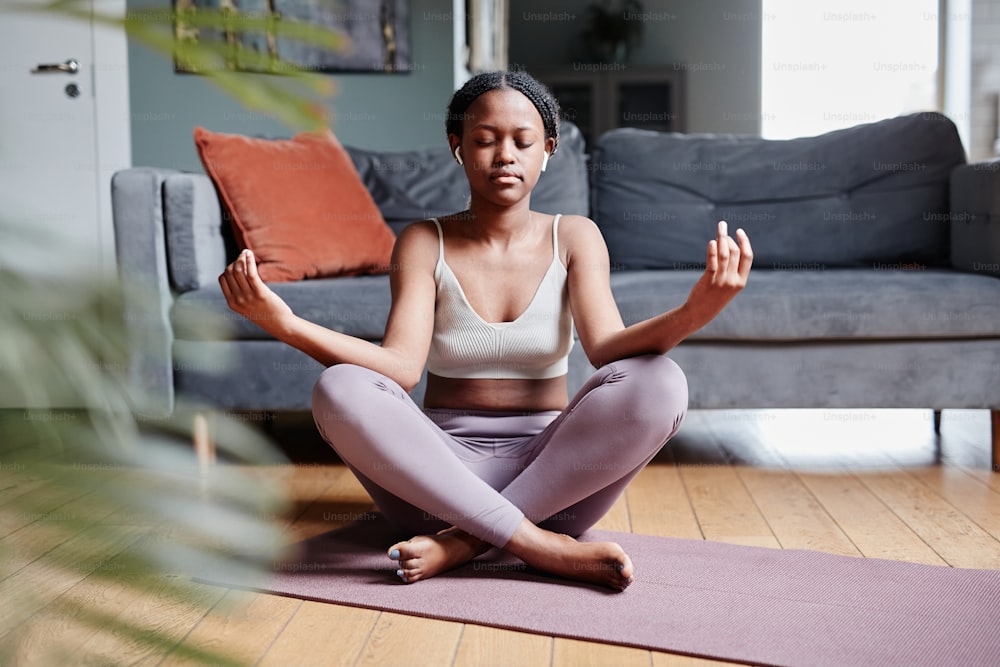 Full length portrait of young African-American woman doing yoga at home while sitting in lotus position with eyes closed