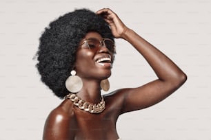Beautiful young African woman in golden jewelry keeping eyes closed and smiling while standing against gray background