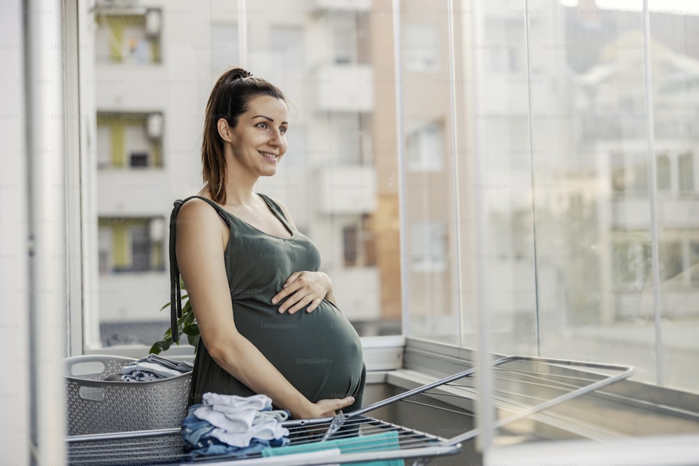 Blessed pregnancy and household chores. A pregnant woman holds her belly while standing on the terrace and looks at the baby clothes that are drying in the tumble dryer. Wife and household chores