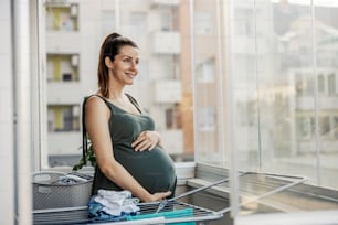 Gesegnete Schwangerschaft und Hausarbeit. Eine schwangere Frau hält sich den Bauch, während sie auf der Terrasse steht und schaut auf die Babykleidung, die im Wäschetrockner trocknet. Ehefrau und Hausarbeit