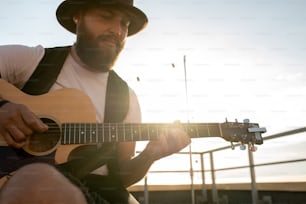 Homem barbudo jovem no chapéu tocando guitarra no telhado em dia ensolarado