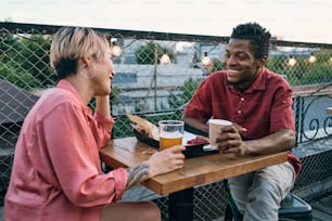 Happy young intercultural couple interacting while having drinks and snacks in outdoor cafe