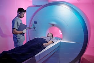 Jovem médico em uniforme pressionando o botão de início da máquina de varredura de ressonância magnética com o paciente deitado em mesa longa