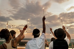 Backs of young intercultural friends with bengal lights raising arms while dancing against cloudy sky